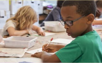 A student in glasses focuses on writing at a classroom table, surrounded by peers who are also engaged in their work. The studious and collaborative atmosphere highlights the importance of advocating for your neurodivergent child at school.